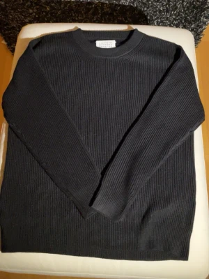 Svart stickad tröja Maison Margiela - Svart ribbstickad tröja från Maison Margiela med klassisk rund hals och långa ärmar. Tröjan har de ikoniska vita stygn bak i nacken och är gjord i mjukt stickat material. Perfekt för dig som gillar minimalistisk och stilren design.