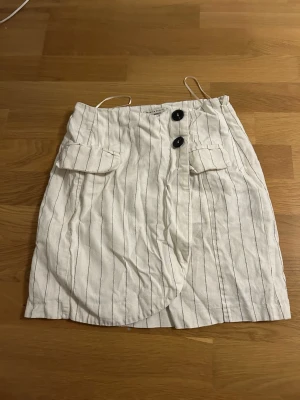 Vit randig kortkjol - Säljer en vit kortkjol från Gina Tricot med svarta tunna ränder. Kjolen har två stora knappar framtill, fickdetaljer och omlott-design. Materialet är bomull och kjolen har en normal passform. Perfekt för dig som gillar stilrena och fräscha plagg.