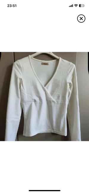 Vit omlottopp från Vero Moda - Säljer en stilren vit omlottopp från Vero Moda med lång ärm och v-ringad hals. Toppen är i mjuk bomull och har en figurnära passform som framhäver siluetten. Perfekt att matcha med jeans eller kjol för en clean look.