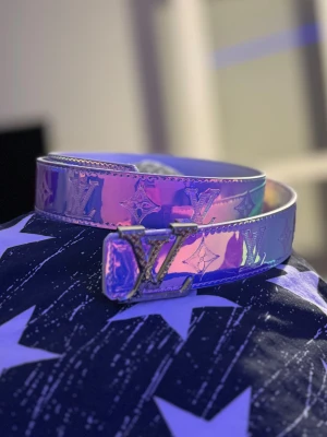 Louis Vuitton holografiskt bälte - Säljer ett Louis Vuitton bälte med holografisk yta i lila och rosa toner. Bältet har klassiskt LV-mönster och en stor LV-spänne i silverfärgad metall. Materialet är syntet med glansig finish och detaljerade sömmar längs kanterna. Perfekt för att lyfta din outfit.