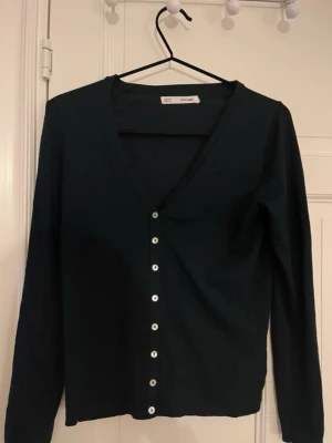 Svart kofta från Sfera Basic XS - Svart kofta från Sfera Basic i storlek XS. Koftan har v-ringning, långa ärmar och vita knappar hela vägen framtill. Enkel och stilren design som passar perfekt till lager-på-lager. Mjuk och skön bomullskvalitet.