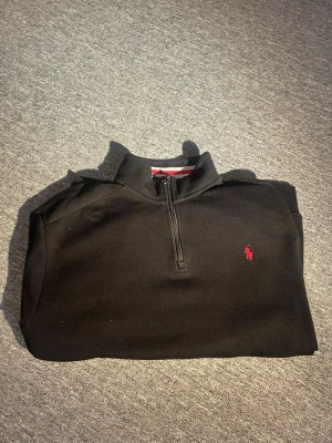 Svart half zip tröja från Polo Ralph Lauren - Svart tröja från Polo Ralph Lauren med röd broderad logga på bröstet. Half zip-dragkedja framtill och ståkrage. Tillverkad i mjuk bomull, perfekt för chill dagar. Snygga ränder på insidan av kragen i rött och vitt.