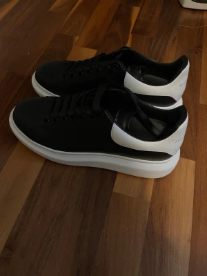 Alexander McQueen svarta och vita sneakers - Säljer ett par stilrena Alexander McQueen sneakers i svart och vitt skinn med chunky vit plattformssula. Skorna har rund tå, svart snörning och logga på både plös och häl. Perfekta för dig som gillar lyxiga och trendiga sneakers med clean design.