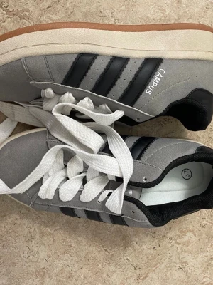 Adidas Campus grå sneakers strl 37 - Säljer ett par Adidas Campus sneakers i grå mocka med svarta ränder och vit sula. Skorna har klassisk snörning med breda vita skosnören och en gummisula i brun färg. Ikonisk retrostil med rund tå och låg profil, perfekt för dig som gillar streetwear.