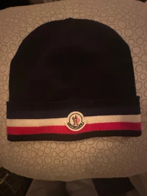 Moncler mössa med röd och vit rand - Snygg mörkblå Moncler mössa med breda ränder i vitt och rött längst ner samt Moncler-logga framtill. Mössan är stickad och har en klassisk form som passar perfekt för kyliga dagar. Perfekt accessoar för dig som gillar stilrena detaljer och exklusiva märken. Lappen finns kvar 