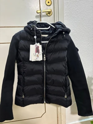 Svart Moncler pufferjacka med huva - Svart pufferjacka från Moncler med huva och dragkedja framtill. Jackan har quiltad kropp i blankt syntetmaterial och stickade svarta ärmar. Fickor med dragkedja och Moncler-logga på ärmen. Perfekt för dig som vill ha en snygg och varm jacka. Trender ute nu! Fick det som julklapps present men hade redan köpt en sen tidigare. Därför säljs den! Den med högst bud får den. Storlek S. Skriv om ni har frågor! ☺️☺️ 