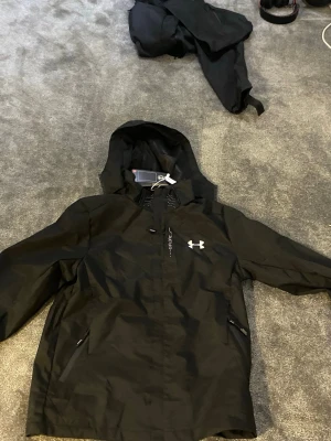 Svart vindjacka från Under Armour - Storlek M men passar mer som S.     Svart vindjacka från Under Armour med huva och dragkedjor på fickorna. Jackan har tryckt logga på bröstet och texten 'Explore' längs dragkedjan. Materialet är lätt och syntetiskt, perfekt för aktiva dagar. Snygg och sportig design med HeatGear-teknologi.
