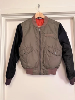 Olivgrön bomberjacka med svarta ärmar - Bomberjacka i olivgrönt med svarta ärmar och orange foder. Jackan har ribbad krage, underkant och muddar, två fickor med lock och knappar samt en ficka med dragkedja på ärmen och en innerficka. Croppad passform. Bra kvalité, som trots flitig användning hållt sig fin. Välomhändertagen men tecken på slitage finns på knapparna till fickorna (se bild). Sälj på flera sidor, först till kvarn. 