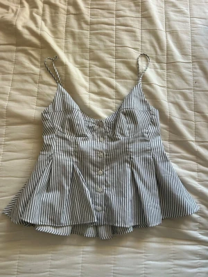 Korsett topp från Zara - Supersöt vit och svart randig linnetopp med knappar framtill och smala axelband i strl xs. Toppen har en lätt utsvängd passform och markerad byst, vilket ger en snygg siluett. Perfekt för varma dagar och enkel att styla med jeans eller kjol.