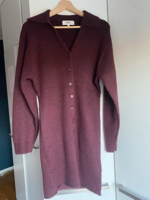 Vinröd stickad långklänning från Gina Tricot - Stickad klänning i ullblandning med knappar framtill från Gina Tricot. Klänningen är vinröd och har intagen midja. Den har v-ringning, lång ärm, figurnära passform och ribbstickning i midjan och vid ärmslut. Använd 1 gång, jättefint skick. 