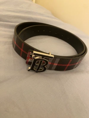 Rutig Burberry skärp i skinn - Säljer ett snyggt Burberry skärp i svart skinn med klassiskt rutigt mönster i grått och rött. Skärpet har en metallspänne med Burberrys ikoniska B-logga och detaljer med riddare. Perfekt för att lyfta din outfit.