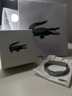 Snygg silverfärgad Lacoste armband - Stilren armband från Lacoste i silverfärgat metallmaterial. Länken har rektangulära länkar med graverad Lacoste-logga på en av länkarna. Kommer med originalpåse och box med klassisk krokodillogo. Perfekt accessoar för att lyfta din outfit.