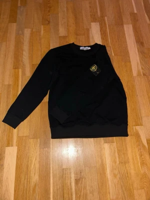 Svart sweatshirt från Stone Island - Säljer en svart sweatshirt från Stone Island med klassisk rund hals och ribbade muddar. Tröjan har lång ärm och den ikoniska Stone Island-loggan på vänster ärm. Liten i storlek. 