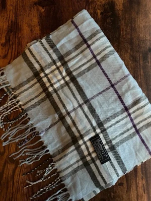 Burberrys Halsduk - Klassisk Burberrys-halsduk i 100% lammull, tillverkad i England. Mjukt och varmt material i ikoniskt rutmönster. Passar både dam och herr. I mycket bra skick utan fläckar eller hål – perfekt för höst och vinter. 