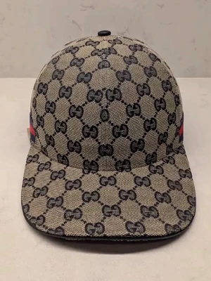 Beige Gucci keps med GG-mönster - Säljer en beige keps från Gucci med det klassiska GG-monogrammet över hela kepsen. Kepsen har justerbart spänne i svart skinn baktill och insida i mörkblått tyg. Snygg och tidlös accessoar för dig som gillar lyxiga detaljer.