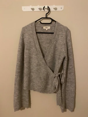 Grå omlottkofta med knytband - Mysig grå stickad omlottkofta från Gina Tricot med knytband i sidan och v-ringad hals. Koftan har lång ärm och är perfekt för lager-på-lager. Enkel att styla till både jeans och kjol.