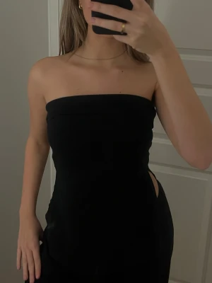 Svart bandeau tubtopp med slits - Säljer en svart bandeau tubtopp med slits i sidan. Toppen är axelbandslös och har en clean, minimalistisk look som passar perfekt till många olika outfits. Materialet känns mjukt och följsamt mot kroppen.