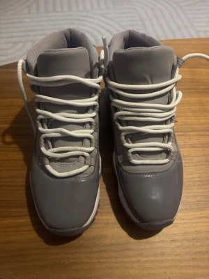 Gråa Nike Jordan 11 sneakers - Säljer ett par gråa Nike Jordan 11 sneakers med vita snören och vit/grå sula. Skorna har hög profil och klassisk rund tå. Materialmix av skinn och syntet med glansig finish på tån. Perfekta för dig som vill ha en ikonisk och stilren sneaker. Lite defekt på tån som enkelt går att fixa. Storlek 42 