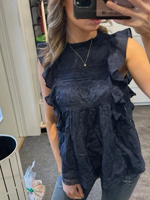 Mörkblå blus med volangdetaljer - Supersöt mörkblå blus med volanger längs axlarna och framtill. Blusen är ärmlös och har en hög halsringning. Tillverkad i ett lätt och luftigt tyg, perfekt för varmare dagar. Passar snyggt till både jeans och kjol för en trendig look.