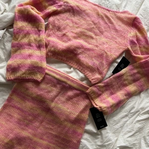 Randig croppad kofta från NA-KD - Supersöt stickat set från NA-KD i rosa och ljusgula ränder. Tröjan är stickad, har lång ärm och twistad omlottdetalj bak. Perfekt för dig som vill ha en färgglad och trendig look. Materialet är mjukt och luftigt. Tröjan är i storlek M ock kjolen i S.