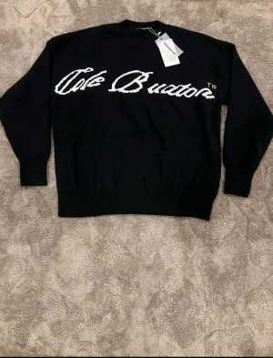Svart stickad tröja Cole - Säljer en svart stickad tröja med vit logga i stor text över bröstet. Tröjan har rund halsringning, ribbade muddar och lång ärm. Perfekt för dig som gillar streetwear och vill ha något stilrent men ändå med statement.