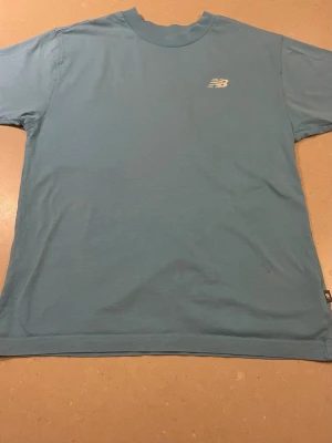 Blå New Balance t-shirt i bomull - Säljer en blå t-shirt från New Balance i storlek S. T-shirten har klassisk passform, korta ärmar och rund hals. Liten NB-logga framtill och stor New Balance-tryck på ryggen. Tillverkad i mjuk bomull, perfekt för en chill och sportig look.