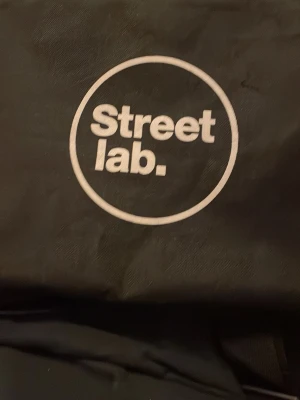 Svart Streetlab skateboard väska - Säljer en svart axelväska från Streetlab med vitt tryck på framsidan. Väskan har en lång, justerbar axelrem och är tillverkad i slitstark polyester. Perfekt för att bära skateboard eller annan utrustning till träningen eller på stan.