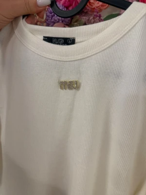 Vit ribbad t-shirt från NLG - Säljer en vit ribbad t-shirt från NLG med ett glittrigt MIU-emblem framtill. T-shirten har klassisk rund halsringning och är tillverkad i mjukt material som känns skönt mot huden. Perfekt för dig som gillar stilrena detaljer.