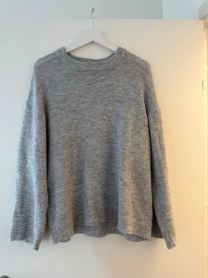 Grå stickad tröja från 157 - Mysig grå stickad tröja från 157 med rund halsringning och avslappnad passform. Perfekt för kyliga dagar och enkel att matcha med jeans eller kjol. Tröjan har ribbade muddar vid ärmslut och nederkant för extra komfort.