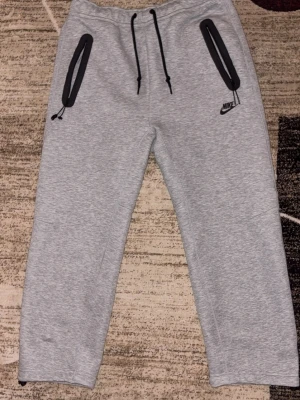 Grå mjukisbyxor från Nike strl L - Använt max 2 gånger. Säljer ett par grå mjukisbyxor från Nike i storlek L. Byxorna har svarta dragkedjor på fickorna, snörning i midjan och Nike-logga på vänster ben. Tillverkade i mjukt bomullsmaterial för maximal komfort. Perfekta för chill eller träning.