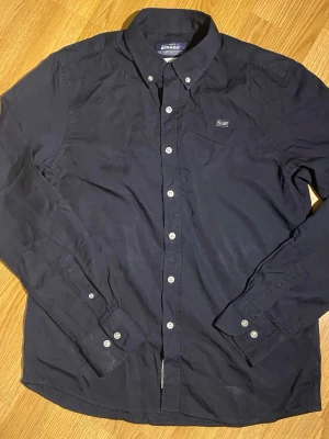 Mörkblå skjorta från Sebago - Klassisk mörkblå skjorta från Sebago med button-down krage och vita knappar. Skjortan har en liten bröstficka med diskret logga och är tillverkad i mjuk bomull. Perfekt till jeans eller chinos för en clean look.