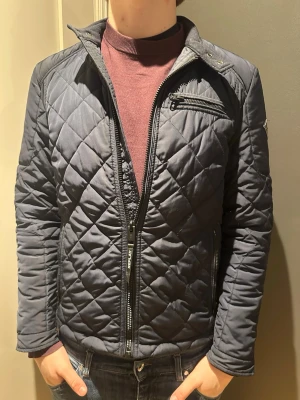 Mörkblå quiltad jacka från Replay - Säljer en mörkblå quiltad jacka från Replay med dragkedja framtill och fickor med dragkedja. Jackan har ett rutigt mönster, lång ärm och Replay-logga på ena ärmen. Perfekt för dig som vill ha en stilren och bekväm jacka till höst och vår.