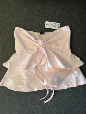 Ljusrosa bandeau tubtopp med volang - Supersöt ljusrosa bandeau tubtopp från H&M med volangdetaljer och snörning framtill. Toppen är axelbandslös och har en mjuk, luftig känsla i bomullsmaterial. Perfekt för sommarens alla tillfällen!