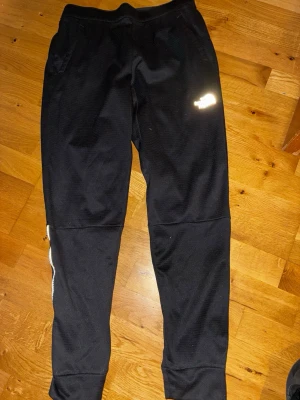 Svarta mjukisbyxor från The North Face - Svarta mjukisbyxor från The North Face med resår i midjan och diskret logga på låret. Tillverkade i mjukt och bekvämt material, perfekta för chill eller träning. Smal passform med muddar vid bensluten och fickor på sidorna.