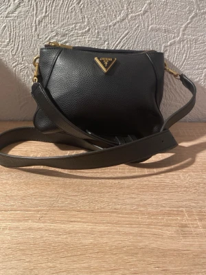 Svart axelväska från Guess i skinn - Snygg svart axelväska från Guess i strukturerat skinn med guldfärgade detaljer och dubbla dragkedjor. Väskan har justerbar rem och invändigt ljust foder med Guess-logga. Perfekt storlek för det viktigaste och stilren design.