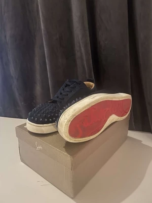Louboutin - Säljer ett par svarta sneakers från Louboutin med ikoniska röda sulor och coola nitar över hela tån. Skorna har snörning, rund tå och vit sula. Utsidan är i textil och insidan i beige skinn för extra komfort. Perfekta för dig som vill sticka ut.