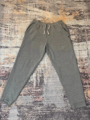 Grå mjukisbyxor från Polo Ralph Lauren XL - Grå mjukisbyxor från Polo Ralph Lauren med dragsko i midjan och broderad logga på vänster sida. Byxorna har ribbade muddar vid bensluten och är tillverkade i mjukt bomullsmaterial. Perfekta för chill dagar eller när du vill vara extra bekväm.