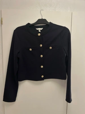 Svart cardigan kofta/jacka från H&M - Snygg svart cardigan jacka från H&M med guldknappar framtill och dekorativa fickor. Jackan har en croppad passform och är tillverkad i ett mjukt material av bomull och polyester. Använt 1 gång.