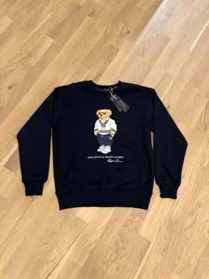 Svart sweatshirt Polo Bear Ralph Lauren - Svart sweatshirt från Polo Ralph Lauren med ikoniskt Polo Bear-tryck på bröstet. Tröjan har rund hals, ribbade muddar och är långärmad. Perfekt för dig som gillar streetstyle och klassiska märken.