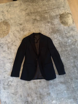 Mörkblå kavaj från Riley - Snygg mörkblå kavaj med två knappar framtill och klassiska fickor. Kavajen har smal krage och helfodrad insida, perfekt för en stilren look. Passar till både skjorta och t-shirt för en clean outfit.