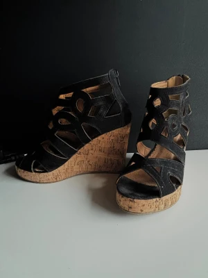 Svarta kilklackade sandaler i mocka - Säljer ett par svarta sandaler med hög kilklack i kork och snygga utskurna detaljer. Ovandelen är i mocka, platån i kork ochdragkedja bak. Perfekta för dig som vill ha både höjd och komfort i sommar. Endast använda en gång