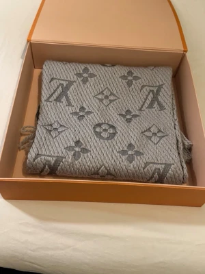 Grå Louis Vuitton stickad halsduk  - Säljer en grå stickad halsduk från Louis Vuitton med det klassiska monogrammönstret i mörkare grå toner. Halsduken har en mjuk och grovstickad känsla med fransar i nederkant. Perfekt för dig som vill ha en lyxig och stilren look.