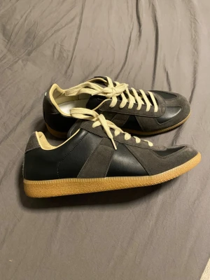 Maison Margiela Gats sneakers svart/grå - Maison Margiela Gat sneakers i svart och grå mocka och skinn, med beige detaljer och snörning. Klassisk gummisula i ljusbrun färg och rund tå. Tillverkade i Italien, riktigt clean och tidlös modell som funkar till det mesta.