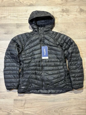 Haglöfs  - Säljer en svart, lätt och riktigt varm dunjacka från Haglöfs, modell Essens Down Hood Women. Jackan har huva, dragkedja framtill och är tillverkad i Pertex Quantum-material med vattenavvisande HyperDRY-dun. Perfekt för kalla dagar och outdoor-äventyr.