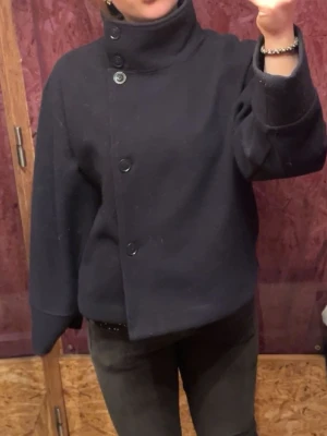  Blå kort kappa  - Snygg mörk blå  jacka i ullblandning med rak, boxig passform och stora knappar framtill. Jackan har höga muddar, lång ärm och finns både med hög krage och klassisk krage. Perfekt för dig som gillar minimalistisk stil och vill ha något som sticker ut lite extra.