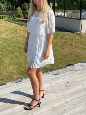 Vit kort sommarklänning med spets  - Säljer en superfin vit kortklänning med plisserad kjol och överdel. Klänningen har korta ärmar och en spetsdetalj vid halsringningen. Perfekt för sommarens alla tillfällen 🌸 Stretchig i midjan. Jag är 163 cm. Publicerad på flera säljsidor. 