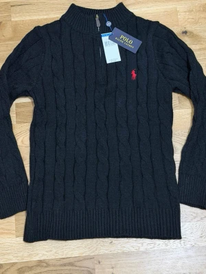Svart Polo Ralph Lauren - Kabelstickad svart tröja från Polo Ralph Lauren med halv dragkedja och röd broderad logga på bröstet. Tröjan har hög krage och långa ärmar, perfekt för kyliga dagar. Klassisk och stilren design som funkar till allt. TVEKA INTE ATT SKICKA EN BUD.
