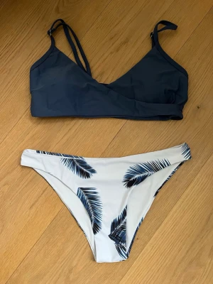Mörkblå bikini från SHEIN - Säljer en snygg bikini från SHEIN strl: M men passar absolut en S, mörkblå topp och vita trosor med blå palmbladsmönster. Bikinitoppen har smala axelband och enkel design, medan underdelen har ett tropiskt mönster.