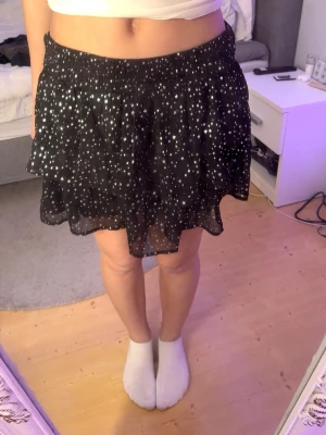 Svart kortkjol med stjärnmönster Kappahl - Supersöt svart kortkjol från Kappahl med vita och silvriga stjärnor över hela tyget. Kjolen har resår i midjan och dubbla volanger för en luftig känsla. Perfekt för dig som gillar en lekfull och trendig stil.