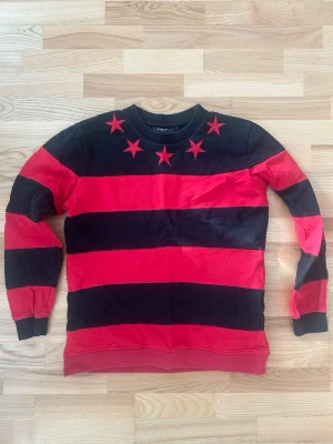 Givenchy randig sweatshirt med stjärnor - Säljer en fet Givenchy sweatshirt i svart och rött med breda ränder och broderade röda stjärnor runt halsen. Tröjan har rund hals, långa ärmar och är gjord i mjuk bomull. Perfekt statement-plagg för dig som vill sticka ut.